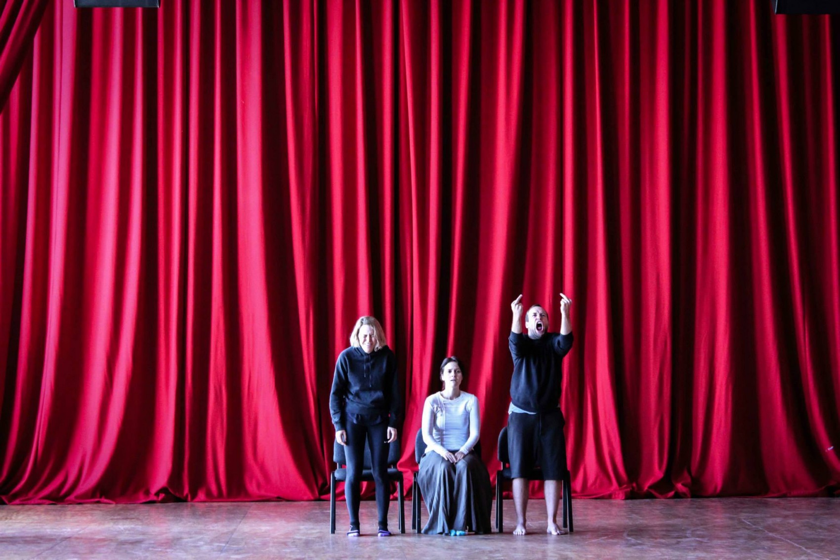 Two standing and one seated performers in a space in front a large red curtain