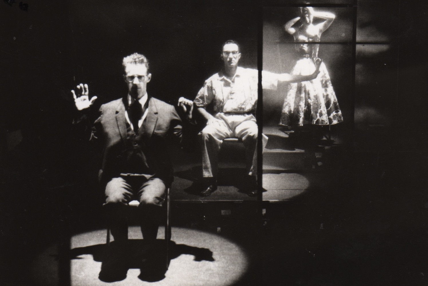 Production image of two men seated in spotlights and one woman standing, arms raised