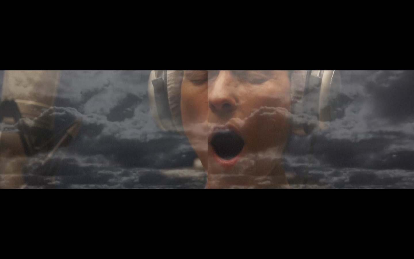 Close shot of woman singing, wearing headphones, behind projection of cloudy sky