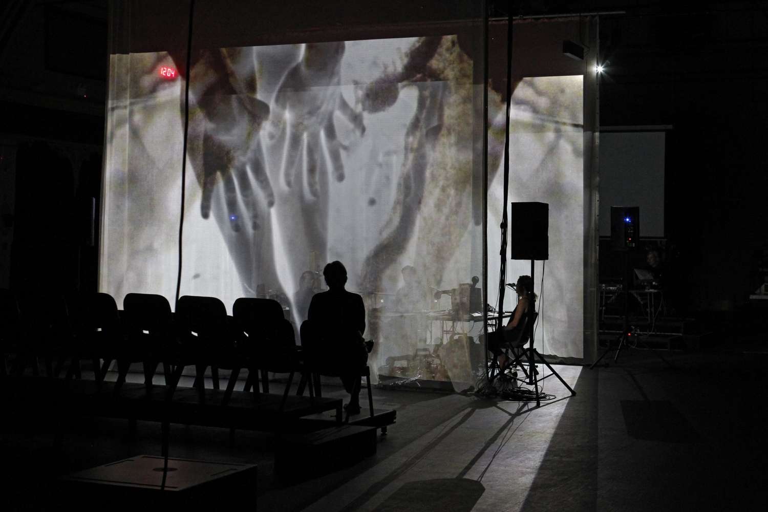 Shot of empty seating bank and two performers seated with large screens on which images of hands are projected