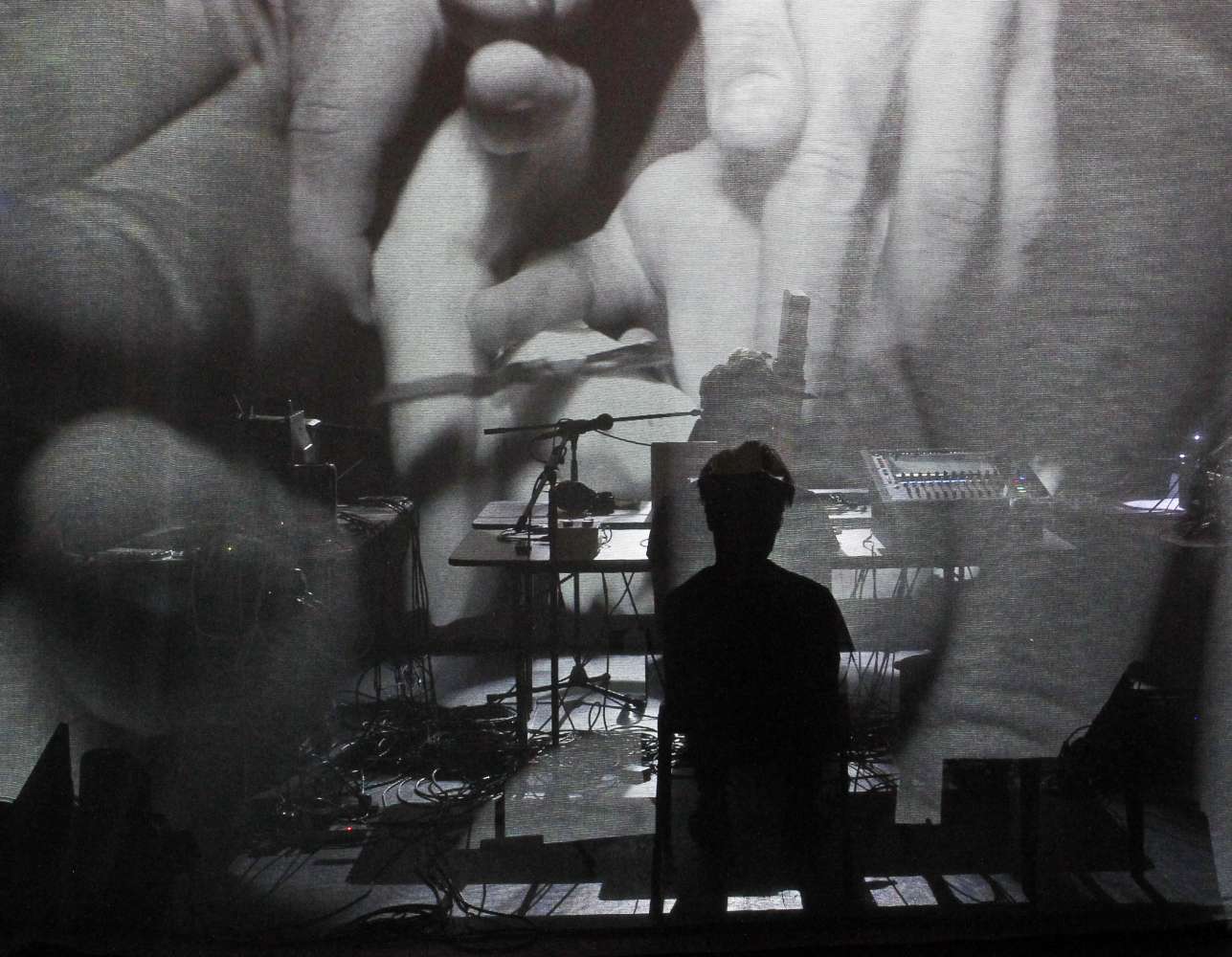Image of a performer and equipment against a large screen projection of intertwined hands