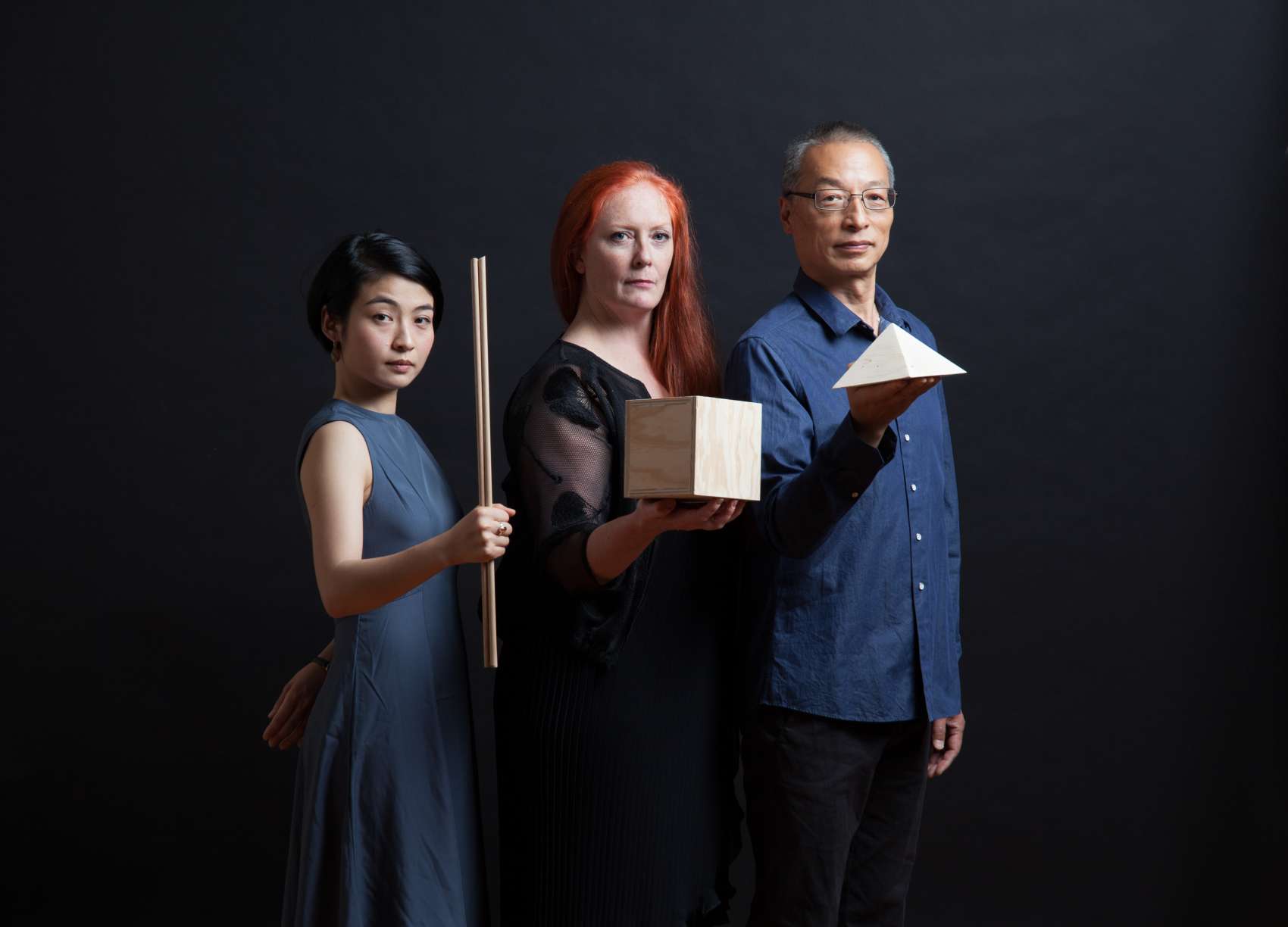 Three performers holding wooden objects standing against a black background