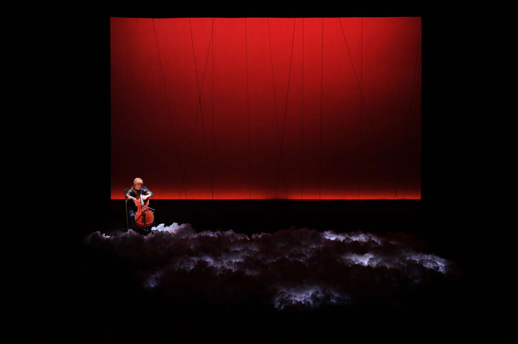 A woman with red hair sits in front of a glowing red screen, playing her cello.On the floor in front of her are clouds.